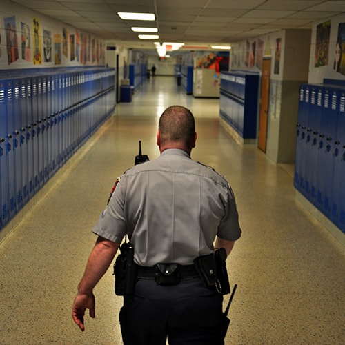 Police officer walking down a school hallway with blue lockers on either side.
