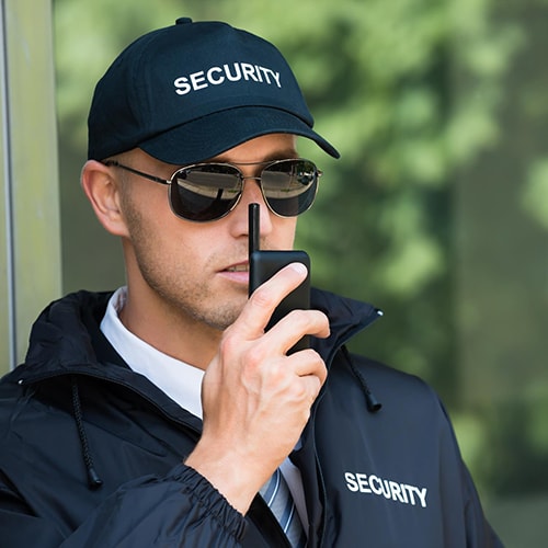 Security guard wearing a cap and jacket with 'SECURITY' on it, using a walkie-talkie.